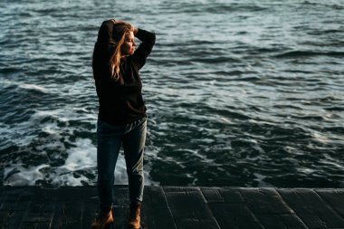  Rıhtımda, deniz kenarında genç ve güzel bir kızın portresi. Şafağa siyah ceket, kot pantolon ve kırmızı ayakkabılarla bakıyor. Ellerinizi başınızın üstüne koyun. Barış, özgürlük, hayatın tatmini