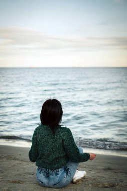  Yeşil örgü süveter ve kot pantolonlu bir kadın deniz kenarında meditasyon yapıyor. Yoga. Düşünen İç Denge