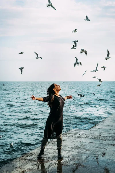 The girl in black stands on the pier with open arms to the sun and the wind receives inspiration and strength and inner peace of the soul seagulls. Open to the challenges and opportunities of life