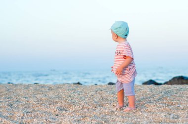 Kumun üzerinde duran ve denize yakın mavi şapka ve çizgili kıyafetler içinde seyir sırt gülümseyen bebek çocuk. Yaz konsepti. Tatil rahatlatıcı, beach tatil.