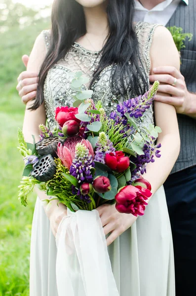 Wedding bouquet with green, red and violet flowers and white ribbon. Bride and groom with wedding bouquet in her hands. Bouquet consists of lupine, peony, protea, eucalyptus and lotus.