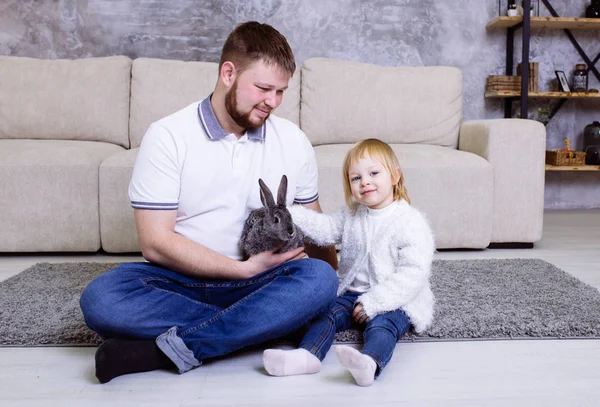 Girl with grey Easter rabbit, bunny and her father. Family Easter celebration. Blonde cute little girl smiling with her father sitting at home on the carpet near the sofa