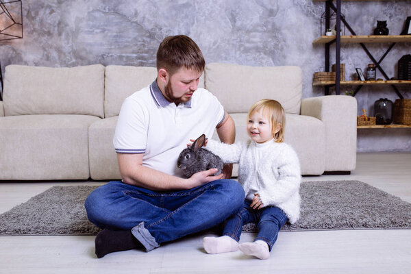 Девочка с серым пасхальным кроликом, кроликом и ее отцом. Семейное празднование Пасхи. Блондинка милая маленькая девочка улыбается, а ее отец сидит дома на ковре рядом с диваном
