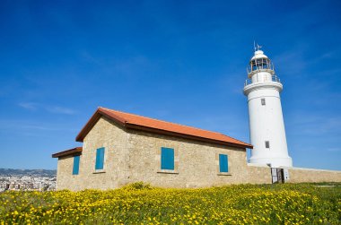 Mavi gökyüzü arka plan üzerinde beyaz deniz feneri. Yaz, deniz ve umut kavramı. Kırmızı çatı kiremitleri ve mavi pencere ile bina taş. Sarı çiçekler ile yeşil alan. Kıbrıs, Paphos.