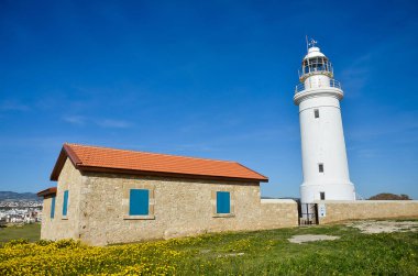 Deniz feneri, Beyaz feneri mavi gökyüzü arka plan üzerinde. Yaz, ilkbahar, deniz ve umut kavramı. Kırmızı çatı kiremitleri ve mavi pencere ile bina taş. Sarı çiçekler ile yeşil alan. yprus, Paphos