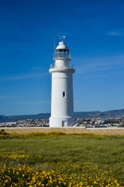 Deniz feneri, Beyaz feneri mavi gökyüzü, dağ ve şehir arka plan üzerinde. Yaz, ilkbahar, deniz ve umut kavramı. Sarı çiçekler ile yeşil alan. Kıbrıs Paphos. Parlak ve renkli