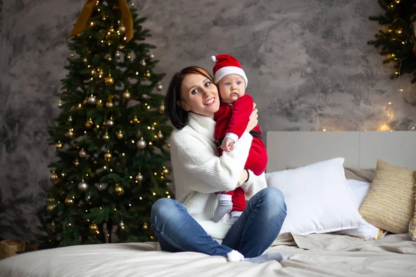 Mère et bébé dans le sourire santa chapeau rouge sur un fond d'arbres de Noël à l'intérieur de la maison — Photo