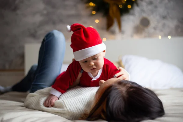 Charmig mor tittar på sitt lilla barn son och ler på bakgrunden av julgranar och kransar i huset — Stockfoto