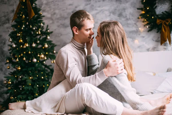 Ζευγάρι ερωτευμένο κάθεται δίπλα σε ένα χριστουγεννιάτικο δέντρο στο κρεβάτι στο σπίτι — Φωτογραφία Αρχείου