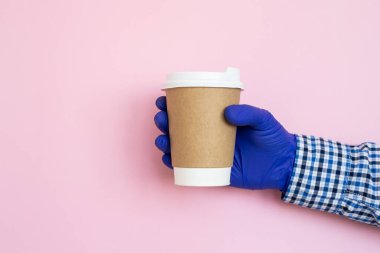 Elinde kahve fincanı ve mavi arka planda tıbbi eldivenler var. Kağıt bardakta kadın eli. Kahve fincanı tutan bir kadın eli. Uzayı kopyala Coronavirus koruması