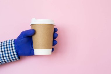 Kahve fincanı tutan bir kadın eli. Uzayı kopyala Elinde kahve fincanı ve mavi arka planda tıbbi eldivenler var. Kağıt bardakla el, koronavirüs koruması.
