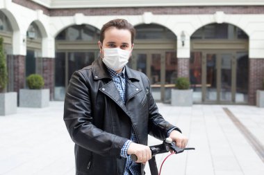 Tıbbi maskeli bir adam modern bir binanın arka planında elektrikli scooter, viral hastalık salgını Coronavirus covid-2019