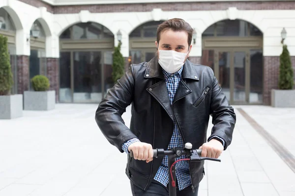 man in a surgical mask on a background of a modern building on electric scooter, coronavirus, illness, infection, quarantine, medical mask