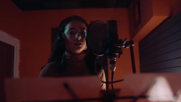 Beautiful young white girl wearing headphones and singing into microphone in recording studio — Stock Video