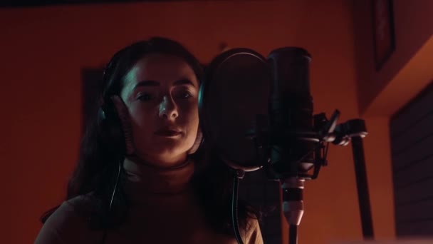 Beautiful young white girl wearing headphones and singing into microphone in recording studio — Stock Video