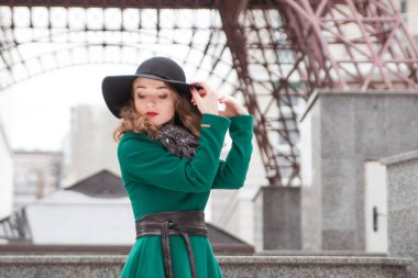 Siyah şapkalı, gri eşarplı, yeşil ceketli bir kız merdivenlerde elinde şapkasıyla dikiliyor..