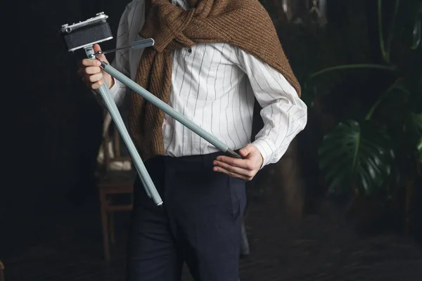 Un hombre con una camisa blanca y un suéter marrón sostiene un trípode viejo con una cámara, un vintage — Foto de Stock