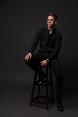 bir adam siyah bir gömlek ve siyah pantolon gri bir arka plan üzerinde koyu bir sandalyede oturur