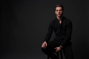 bir adam siyah bir gömlek ve siyah pantolon gri bir arka plan üzerinde koyu bir sandalyede oturur