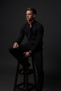 bir adam siyah bir gömlek ve siyah pantolon gri bir arka plan üzerinde koyu bir sandalyede oturur