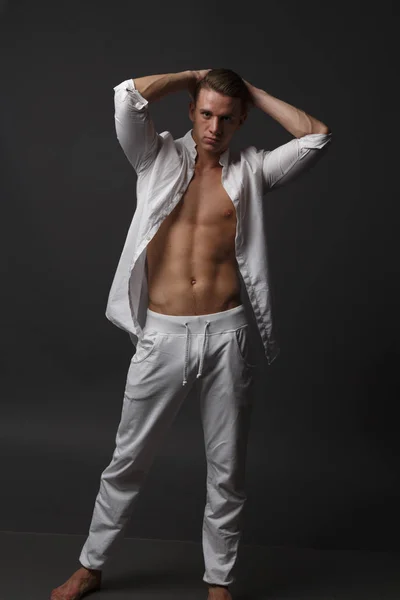 Un mec avec un torse nu, en pantalon blanc, et dans une chemise blanche, se tient sur un fond gris — Photo