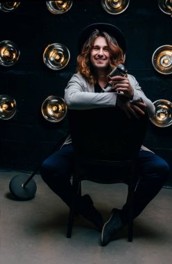 uzun saçlı bir mikrofon, güleryüzlü, ışıklar, arka planı ile onun elinde bir sandalyede oturan adam spot