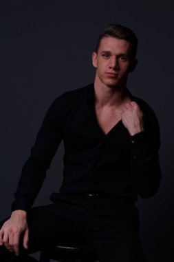 genç, beyaz bir adam siyah bir gömlek ve siyah pantolon, bir sandalyede oturur gömlek siyah bir arka plan karşı düzeltiyor ve kameraya bakıyor. iş, modeli, röportaj, portre