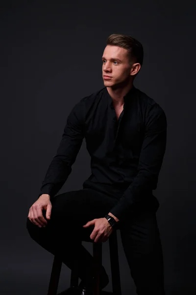 a young, white guy sits on a chair in a black shirt and black pants, against a black background and looking away. business, model, interview, portrait