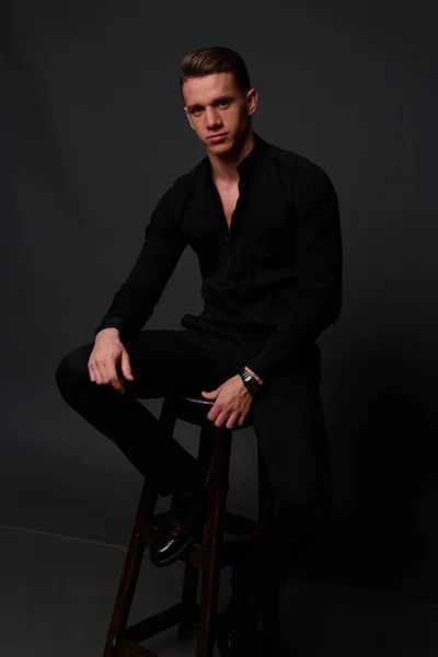 Genç, beyaz bir adam siyah bir gömlek ve siyah pantolon, siyah bir arka plan ve kameraya bakarak karşı sandalyeye oturur. iş, modeli, röportaj, portre — Stok fotoğraf