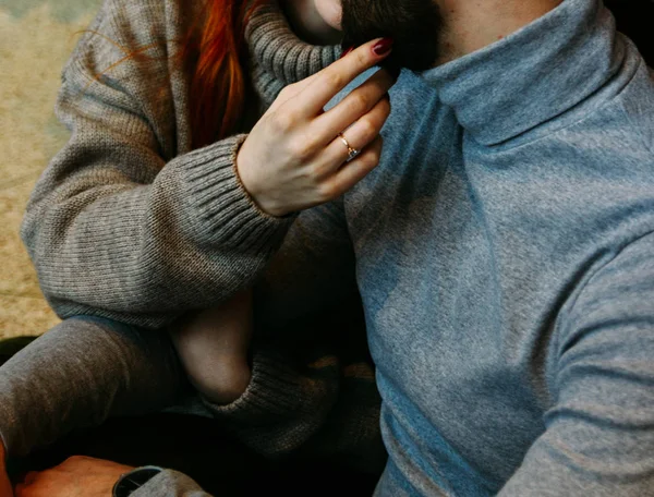 The couple is sitting on the floor in an embrace. embrace. a female hand with a ring stretches to touch the face of a man. kiss talking. gray sweaters. — Stock Photo, Image