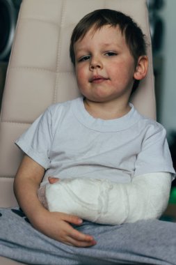 küçük bir çocuk alçıda kırık bir kol ile bir sandalyede oturuyor. hastane. Tıp. tehlike. ağrı. bacaklarda kırmak için. şiddet.