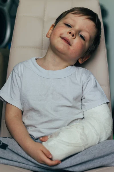 A small boy is sitting on a chair with a broken arm, in a cast. hospital. medicine. danger. pain. to break limbs. violence. — Stock Photo, Image