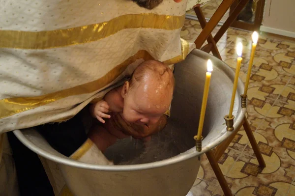 Bautismo en la iglesia, el sacerdote sostiene al niño sobre la fuente y lo sumerge en el agua. el agua fluye sobre la cabeza del niño, el niño llora. queman tres velas . — Foto de Stock