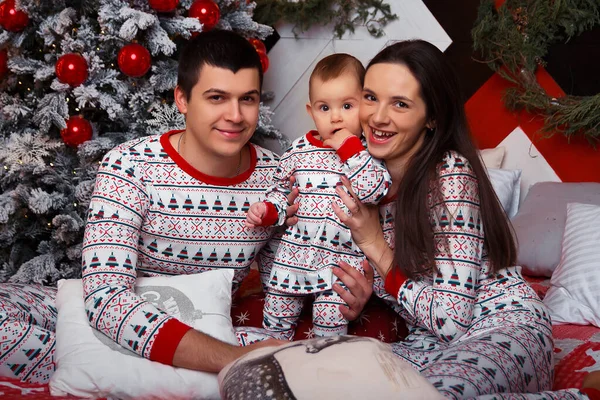 Família em pijama senta-se em uma cama perto da árvore de Natal mãe, pai e criança, idílio, celebração de Natal. felicidade familiar . — Fotografia de Stock