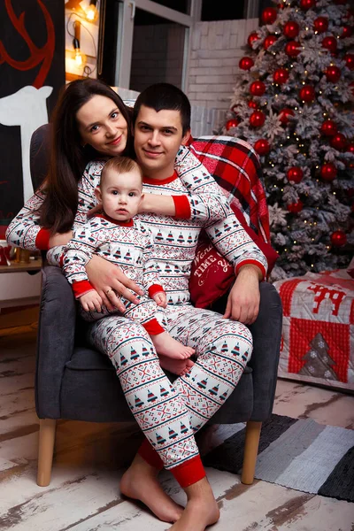 mother hugs her husband from behind, who is sitting on a chair with a baby in her arms, near a Christmas tree in home pajamas