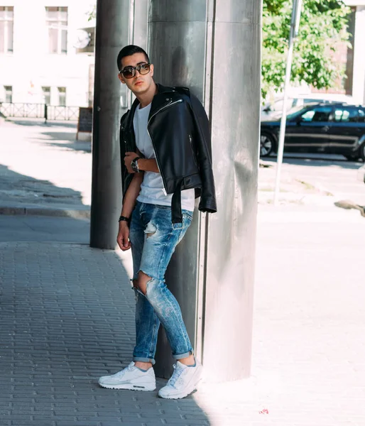 Junger, gutaussehender Typ in weißem T-Shirt, schwarzer Lederjacke und blauer, zerrissener Jeans steht auf der Straße, Modell, Stadtspaziergang, Live-Style. in weißen Turnschuhen — Stockfoto