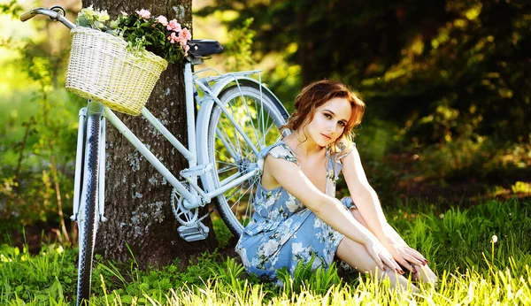 Retrato de una hermosa niña en el bosque, sosteniendo una bicicleta con una cesta de flores, detrás de los rayos del sol, un vestido de flores azules, paseo de verano — Foto de Stock