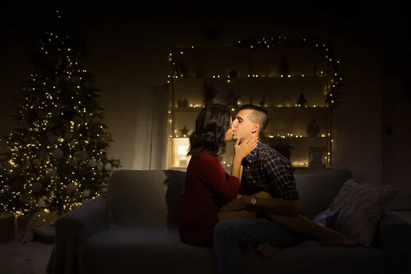 Feliz, casal apaixonado no escuro, noite no sofá, fundo escuro, luzes da árvore de natal. Noite de Natal. Ano Novo. abraço e beijo. atração, noite romântica e data . — Fotografia de Stock