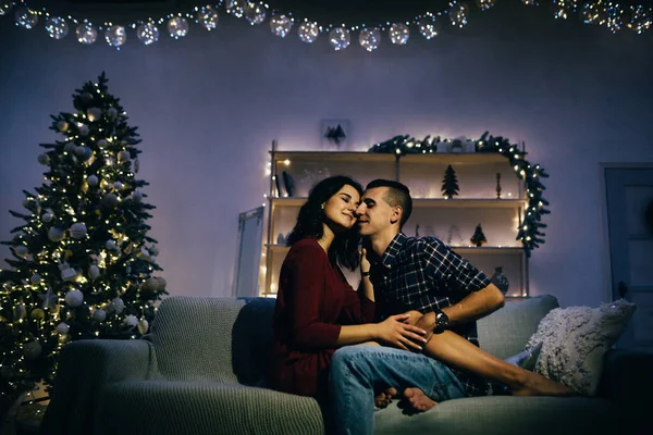 Feliz, casal apaixonado no escuro, noite no sofá, fundo escuro, luzes da árvore de natal. Noite de Natal. Ano Novo. abraço e beijo. atração, noite romântica e data . — Fotografia de Stock