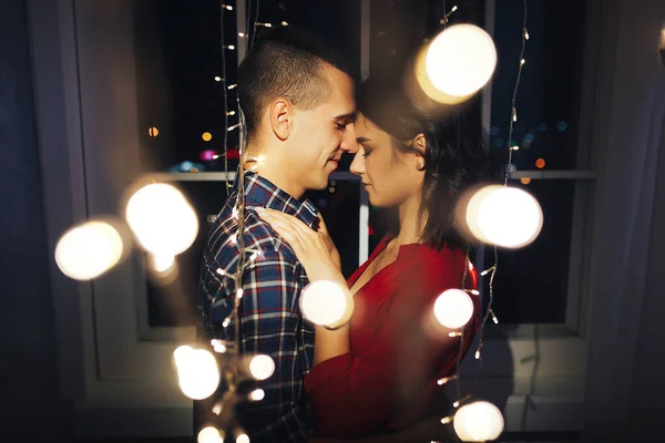 Para zakochana w ciemności pomiędzy światłami girlandy przytula się i całuje. święto i data Bożego Narodzenia. 12 godzin, pocałunek w Sylwestra. w światłach na tle okna. szczęśliwy. — Zdjęcie stockowe