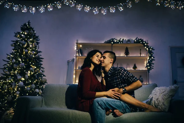 Feliz, casal apaixonado no escuro, noite no sofá, fundo escuro, luzes da árvore de natal. Noite de Natal. Ano Novo. abraço e beijo. atração, noite romântica e data . — Fotografia de Stock