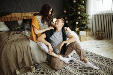 Mutlu çift sabahları Noel ağacının yanındaki yatakta birbirine aşık olur, sarılır, Noel sabahı. Yakınlarda, yerde kahve fincanları var. Ocak, ev kıyafetleri ve kazaklar. konfor ve kalplerin sıcaklığı