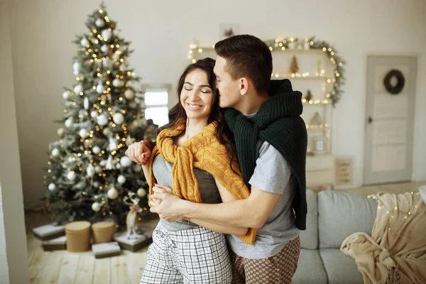 快乐的一对恋人在早上靠近圣诞树，一个男人拥抱一个女孩，早上是圣诞节。 壁炉,家居服和毛衣. — 图库照片