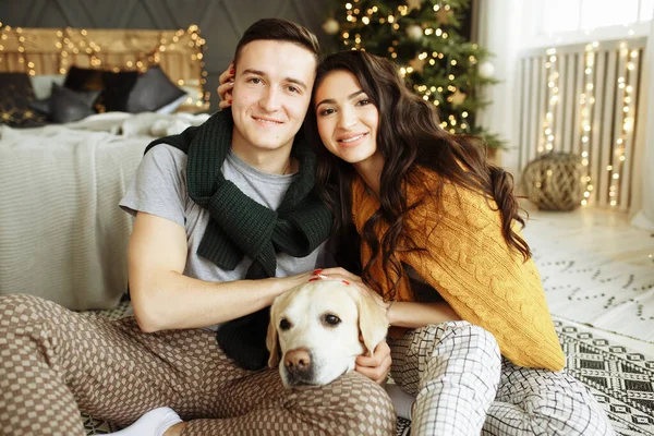 Happy couple is sitting near the bed with a dog, christmas. new year at home. family and date — ストック写真