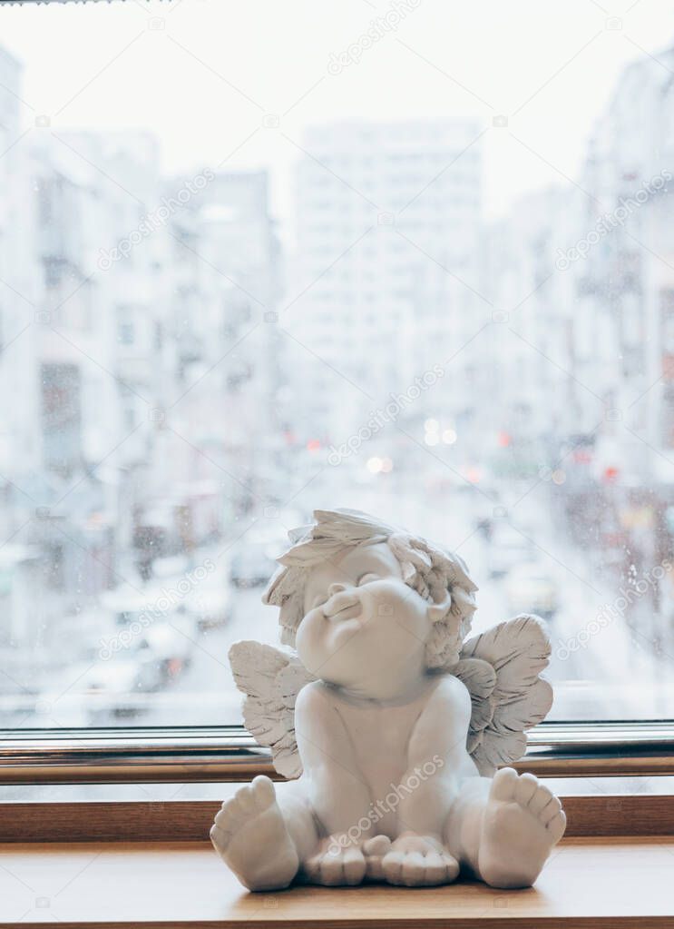 sculpture of an angel, decor, stands on the background of the city, an angel sits on a wooden table, windowsill. Valentine's Day. protection. Guardian