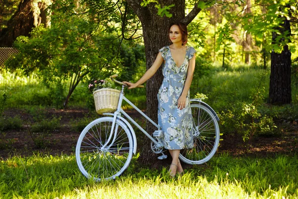 Retrato de una hermosa niña en el bosque, sosteniendo una bicicleta con una cesta de flores, detrás de los rayos del sol, un vestido de flores azules, paseo de verano — Foto de Stock