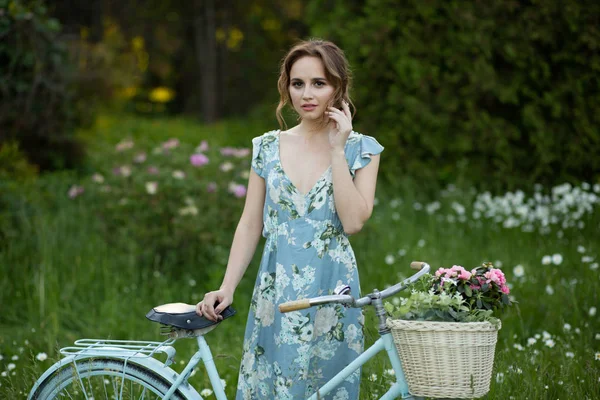 Retrato de uma menina bonita na floresta, segurando uma bicicleta com uma cesta de flores, atrás dos raios do sol, um vestido de flor azul, passeio de verão — Fotografia de Stock