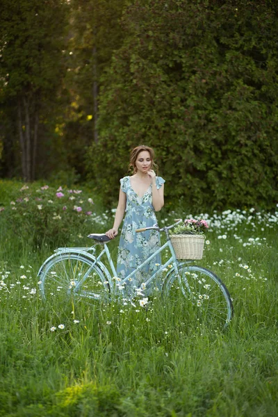 Retrato de uma menina bonita na floresta, segurando uma bicicleta com uma cesta de flores, atrás dos raios do sol, um vestido de flor azul, passeio de verão — Fotografia de Stock