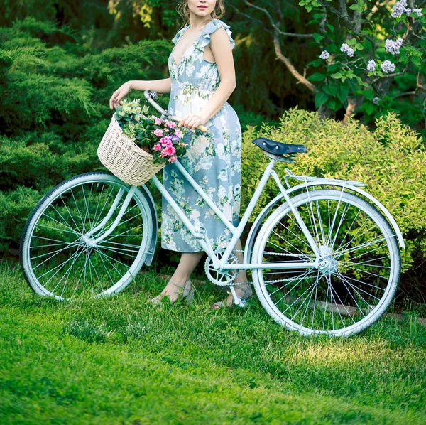 Ritratto di una bella ragazza nel bosco, con in mano una bicicletta con un cesto di fiori, dietro i raggi del sole, un vestito a fiori blu, passeggiata estiva — Foto Stock