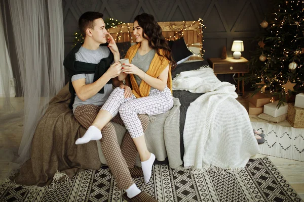 Ευτυχισμένο ζευγάρι ερωτευμένο το πρωί στο κρεβάτι κοντά στο χριστουγεννιάτικο δέντρο, αγκαλιά, το πρωί των Χριστουγέννων. ρούχα του σπιτιού και πουλόβερ. άνεση και ζεστασιά της καρδιάς — Φωτογραφία Αρχείου